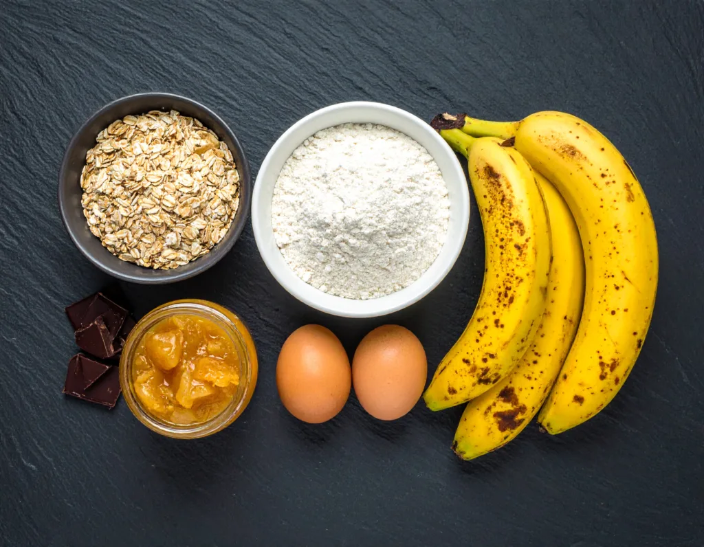 Les ingrédients pour la recette banana bread healthy : bananes mûres, farine complète, compote de pommes et chocolat noir, présentés en vue de dessus.
